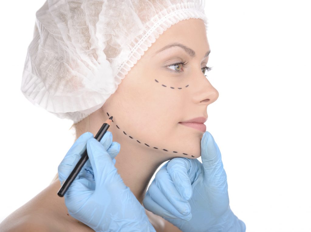 Making marks on face. Close-up of beautiful young woman in medical headwear keeping while doctors hands in gloves making marks on her face isolated on white